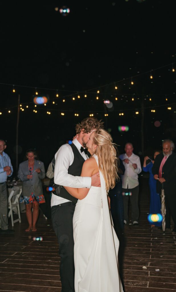 Bride and groom share a final dance under glowing string lights as wedding guests surround them, blowing bubbles during their romantic Cancun resort send-off.