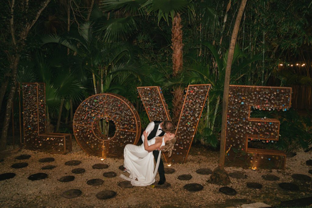 Newlyweds share a dramatic dip kiss in front of a giant LOVE sign covered in locks at their Dreams Riviera Cancun wedding, symbolizing their everlasting love.
