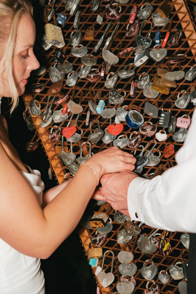 Bride and groom clasp hands as they secure a love lock onto a heart-covered installation, marking their forever at their Cancun destination wedding.