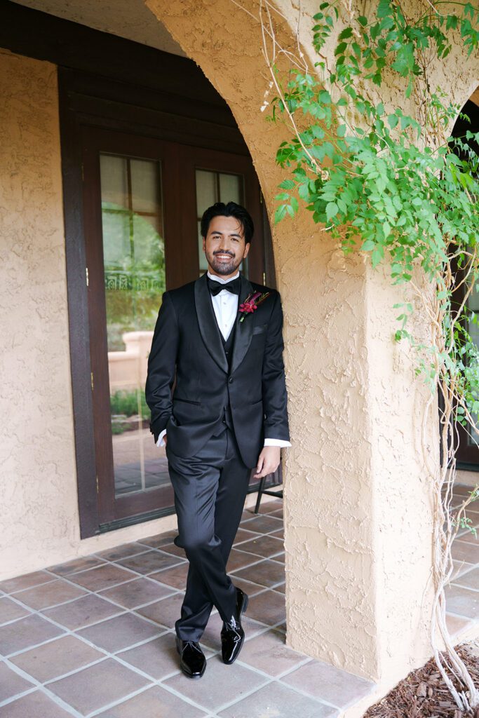 Groom in a black tuxedo standing under a vine-covered archway at Villa Parker
