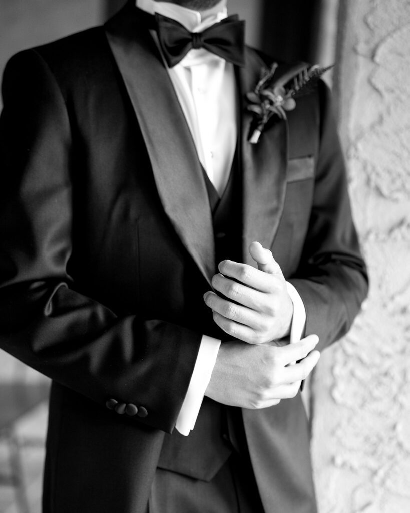 Groom in a black tuxedo with a red boutonniere adjusting his cuff against a stucco wall