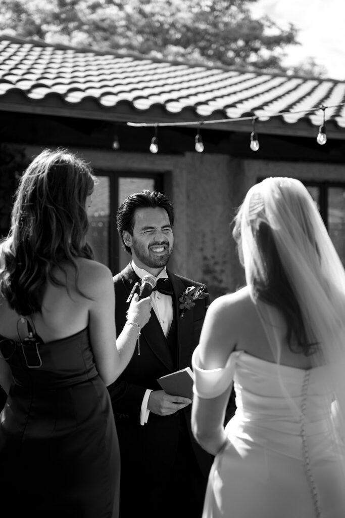 Groom laughing while reading his vows as the officiant holds a microphone