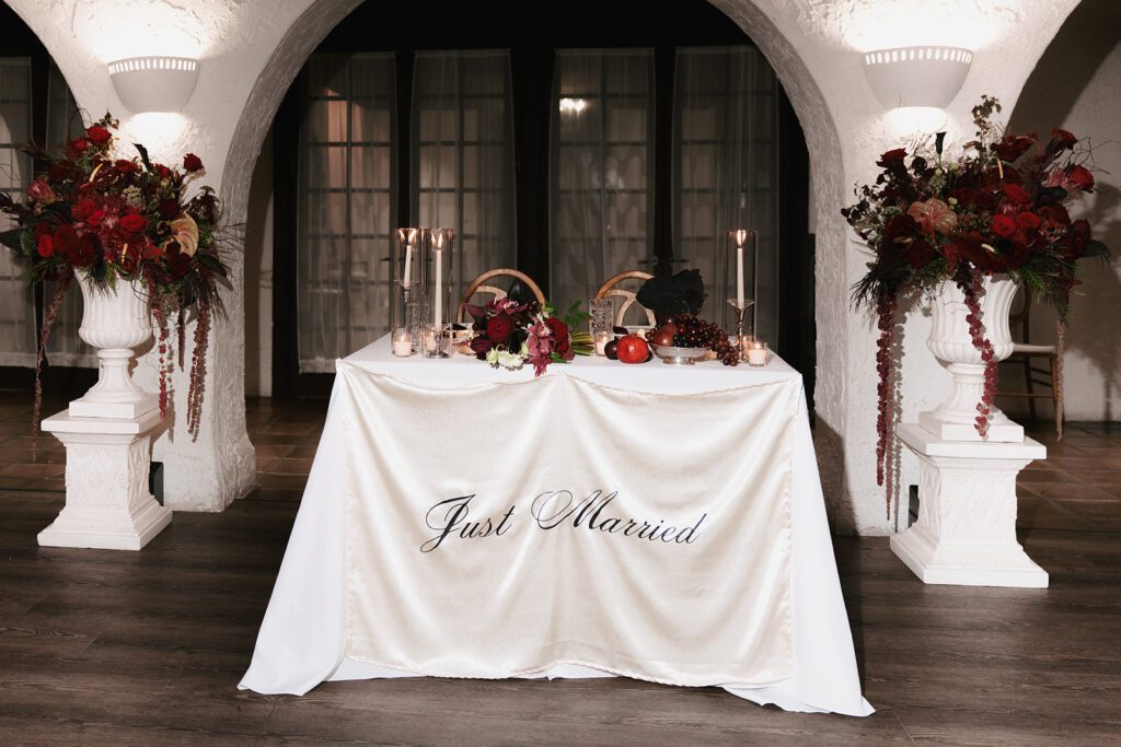 Sweetheart table draped in Just Married satin between tall red floral urns at Villa Parker