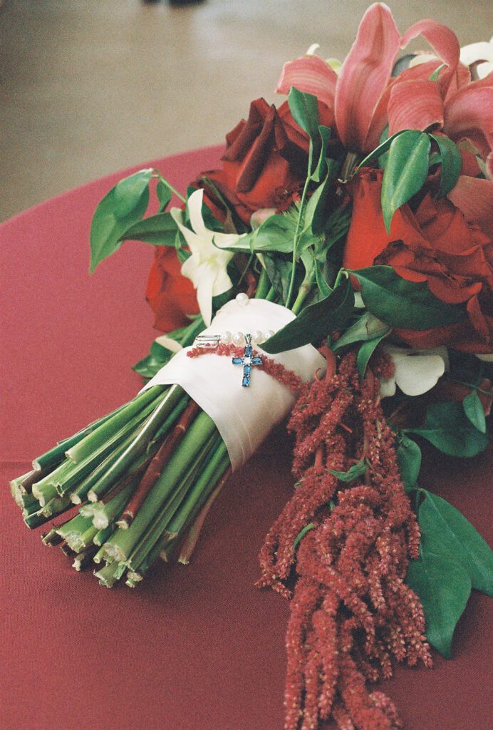 Bridal bouquet of red roses and lilies wrapped in silk with a sapphire cross charm